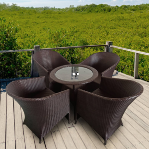 The image shows an outdoor patio setup featuring a round glass-top table surrounded by four dark brown wicker chairs. The furniture is placed on a wooden deck with a railing, overlooking a lush green landscape, possibly a forest or mangrove area, under a bright blue sky with scattered clouds. The setting gives off a relaxing and natural vibe, ideal for outdoor dining or lounging.