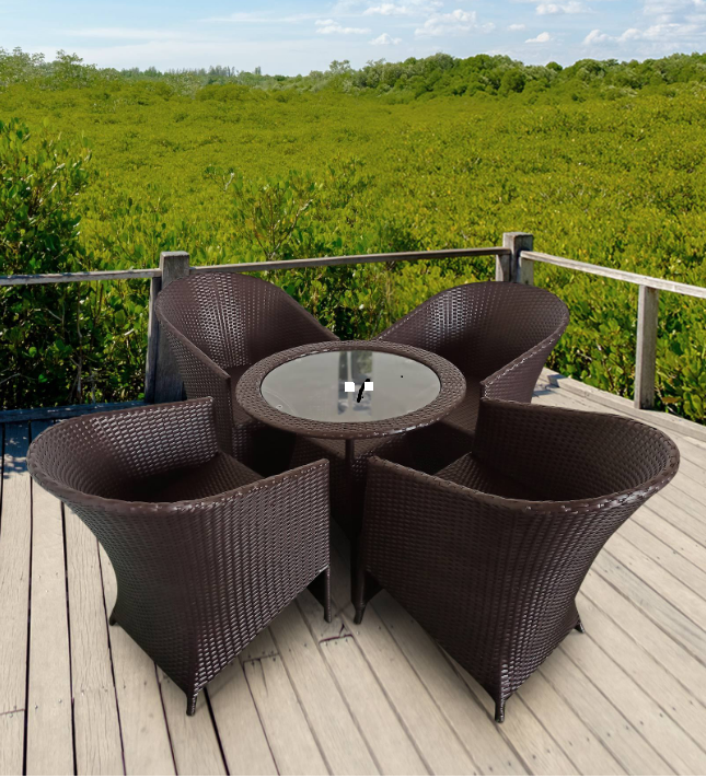 The image shows an outdoor patio setup featuring a round glass-top table surrounded by four dark brown wicker chairs. The furniture is placed on a wooden deck with a railing, overlooking a lush green landscape, possibly a forest or mangrove area, under a bright blue sky with scattered clouds. The setting gives off a relaxing and natural vibe, ideal for outdoor dining or lounging.